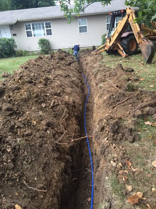 A fresh trench across a front yard with a new blue water line laid in it and a backhoe beyond