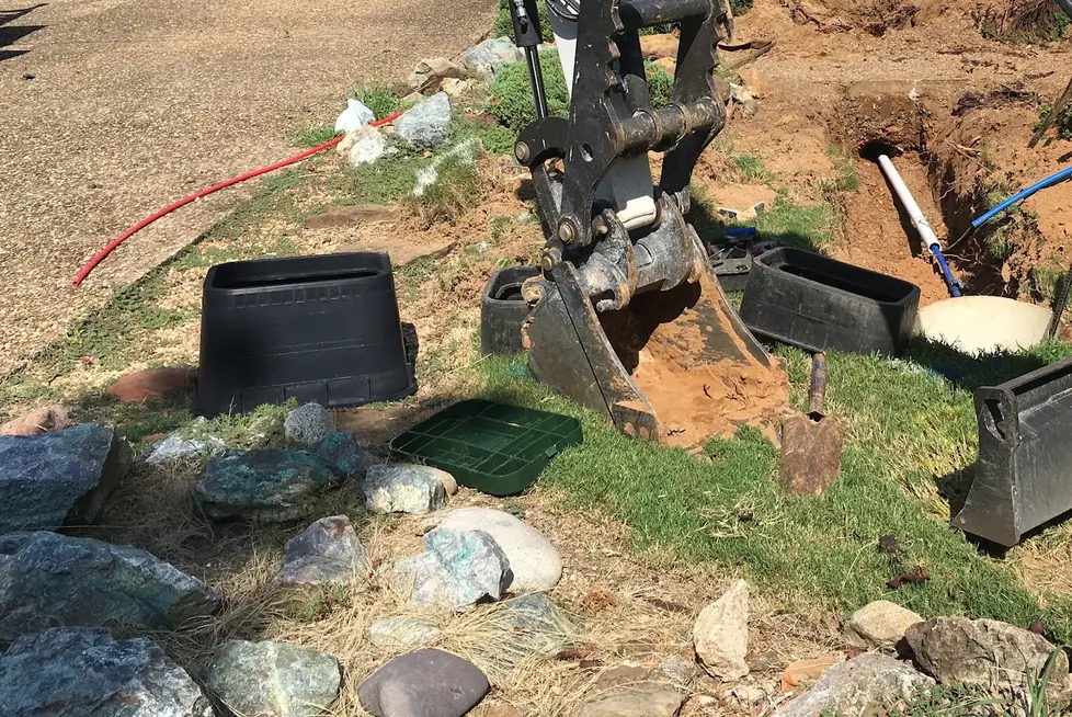 A Hansen yard-line dig: excavator bucket of red clay, two meter boxes and the new line in the open trench