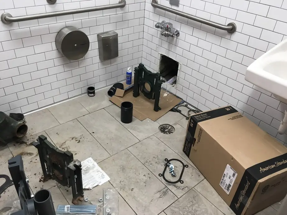 A commercial restroom mid-job with the old toilet carrier removed and a new American Standard toilet boxed