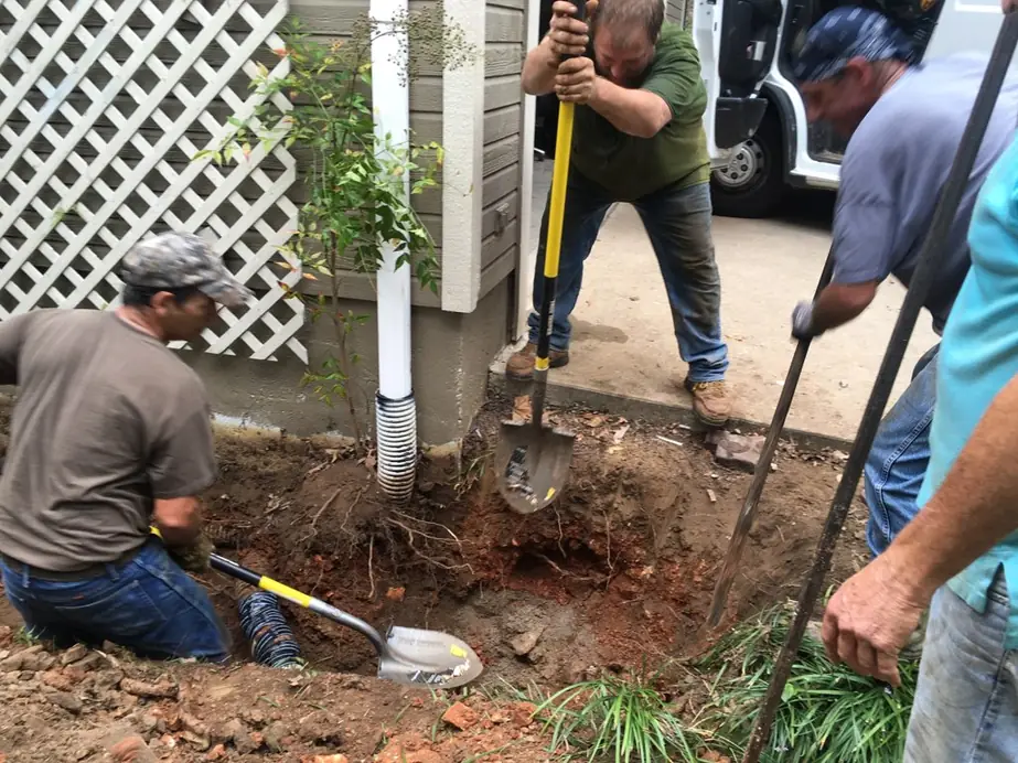 Hansen crew hand-digging around a drain line beside a house, the work van parked behind
